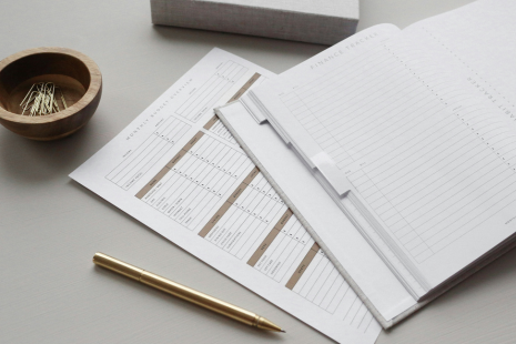 Open planner, paper sheets, golden pen, bowl with paperclips on a desk.